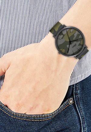 Montre-bracelet avec un motif camouflage vert et gris sur un bracelet noir, portée au poignet gauche, main reposant dans la poche d’un jean bleu.