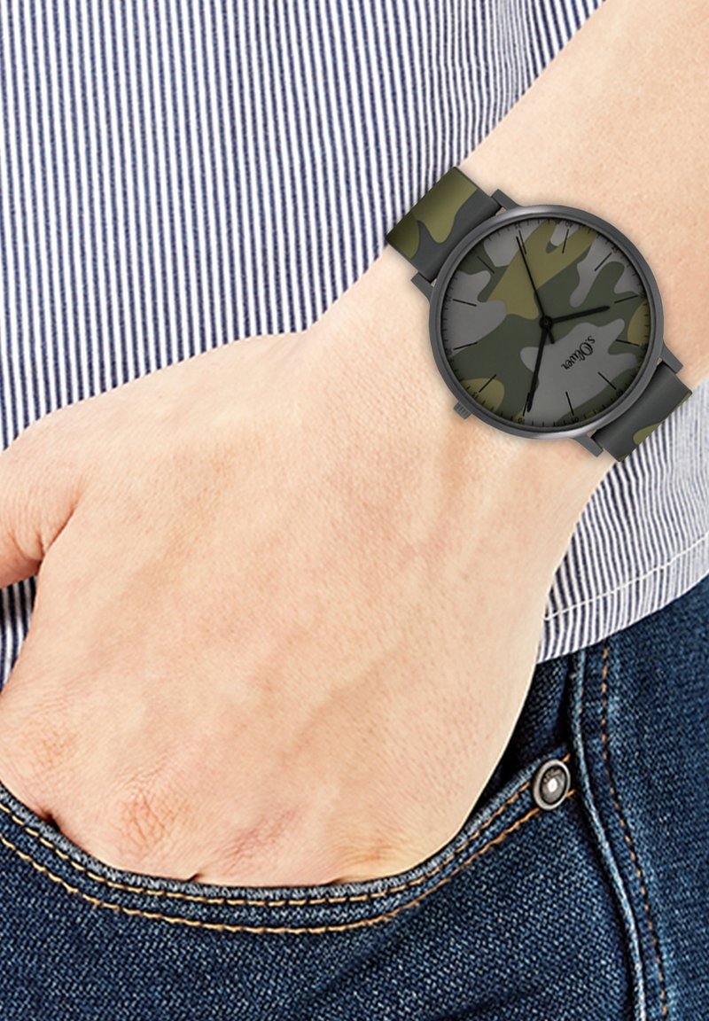 Montre-bracelet avec un motif camouflage vert et gris sur un bracelet noir, portée au poignet gauche, main reposant dans la poche d’un jean bleu.