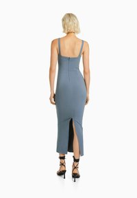 Niebieska sukienka bez rękawów o dopasowanym kroju, do kolan, z bocznym rozcięciem i zapięciem na zamek z tyłu; połączona z czarnymi sandałami na paski.