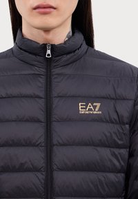 Veste noire matelassée avec col haut et fermeture éclair frontale, marquée EA7 Emporio Armani en or sur le côté gauche de la poitrine, portée par une personne aux longs cheveux foncés.