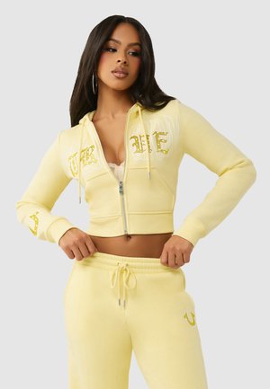 Kobieta dopasowująca sznurek w żółtych spodniach dresowych, ubrana w pasującą krótka bluzę z kapturem zapinaną na zamek oraz koronkowy top pod spodem.