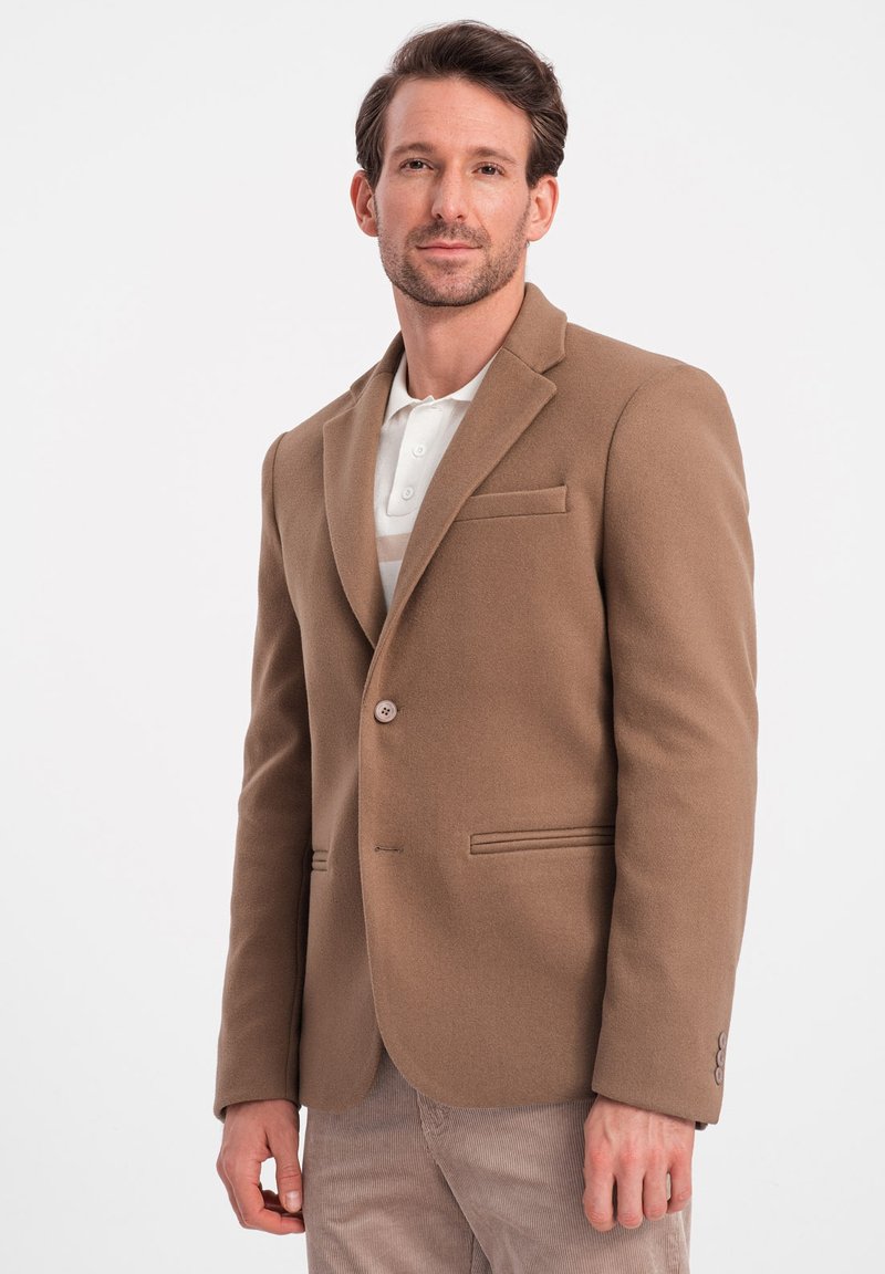Bruine wollen blazer met een getailleerde pasvorm, met een uitgesneden revers, enkele knoopsluiting en een borstzak, gedragen over een wit polohemd.