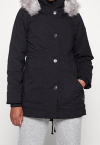 Svart parka-jacka med huva i fuskpäls, med knappstängning, två framfickor och diagonala sömmar på kroppen.