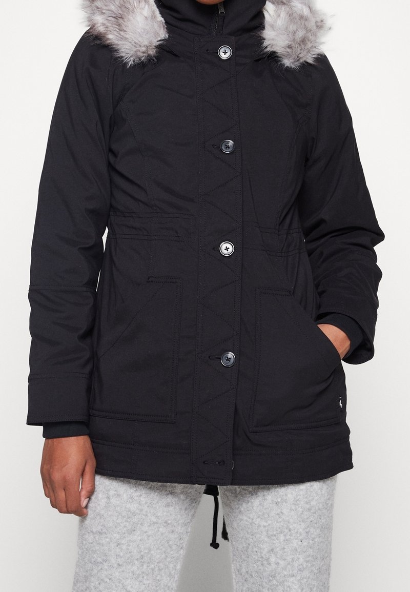 Svart parka-jacka med huva i fuskpäls, med knappstängning, två framfickor och diagonala sömmar på kroppen.