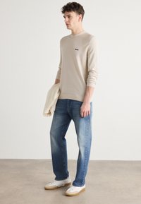 Beige pitkähihaisen paidan, jossa on pieni musta logo, yhdistettynä vaaleansinisiin farkkuihin. Valkoiset tennarit, joissa on kumipohjat. Rentoa, väljä istuvuus.