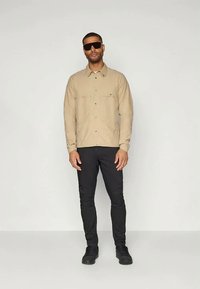 Lysbrun langærmet skjorte med knapper, to brystlommer; parret med sorte bukser og sorte sneakers, båret af en mand i solbriller.