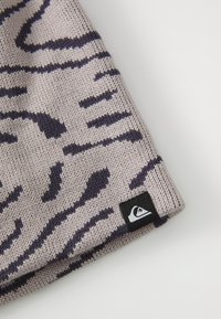 Berretto lavorato a maglia con uno sfondo beige chiaro e un motivo astratto di animali in viola scuro. Presenta un piccolo patch con logo nero sull'orlo.