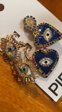 Τρία ζευγάρια σκουλαρίκια από μέταλλο σε χρυσή απόχρωση. Οι σχεδιασμοί περιλαμβάνουν μπλε και πολύχρωμους κρύσταλλους με μοτίβα ματιών και αιχμές.