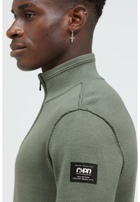 Olivgrüner Sweater mit Reißverschluss, geripptem Kragen, strukturiertem Stoff und Schulternahtdetails. Mit einem schwarzen Logopatch am Ärmel.