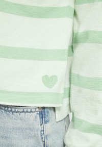 Camicia a maniche lunghe a righe verde chiaro e bianco con tessuto testurizzato, dotata di un'etichetta con logo a forma di cuore sul lato sinistro. Abbinata a jeans in denim.