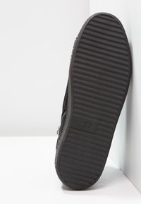Sole of a black shoe with horizontal grooves, size 37 marked on the bottom, positioned vertically against a white wall and floor.
