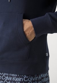 Marineblaue Baumwoll- sweatshirt mit einer Kängurutasche und gerippten Bündchen. Hat ein kleines Logoschildchen nahe dem Saum. Weicher Stoff und klassisches Design.