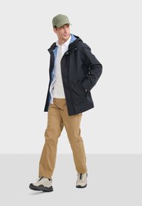 Sort vandtæt jakke med en blå indvendig foring, beige bukser og grå- og sort vandrestøvle, suppleret med en grøn cap.