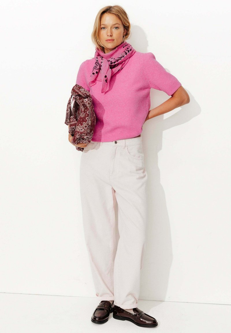 Top en tricot fuchsia à manches courtes, pantalon large côtelé crème, et mocassins marron. Tenant une écharpe à motifs marron avec des accents roses.