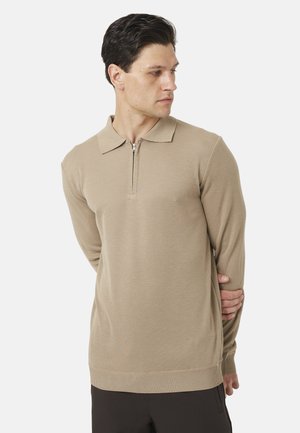 Uomo con capelli scuri che indossa un maglione beige a maniche lunghe con cerniera, guarda verso destra, con una mano che tiene il braccio opposto dietro la schiena.