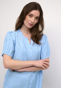 Jasnoniebieska bluzka z krótkim rękawem z zapięciem na guziki z przodu, z marszczonymi detalami na ramionach oraz okrągłym dekoltem. Wykonana z miękkiej, teksturowanej tkaniny.