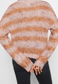 Personne portant un pull en tricot rayé rose et orange, les mains jointes dans le dos, associée à un pantalon noir.