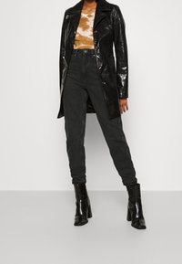 Černý lesklý trenčkot přes top s tie-dye vzorem v hnědé a krémové barvě, doplněný o zúžené tmavé džíny a černé boty na podpatku.