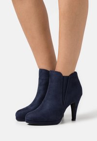 Stivali alla caviglia in suede blu navy con punta affusolata, tacchi alti e sottili, e pannelli laterali elastici per una facile calzata. Texture liscia, dettagli minimi.