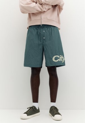 Man die groene geruite korte broek draagt met het woord "City" op zijn linkerbeen, beige jas met ritssluiting, witte sokken en groene sneakers, staand met gekruiste armen.