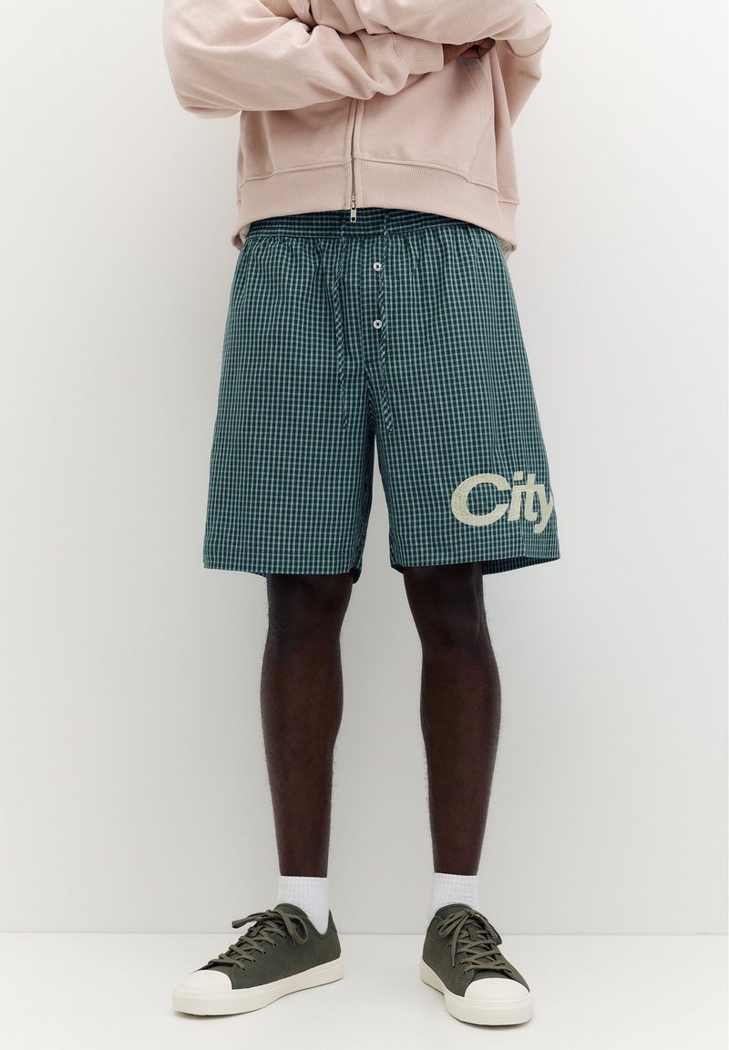 Homme portant un short vert à carreaux avec le mot "City" sur la jambe gauche, une veste beige zippée, des chaussettes blanches et des baskets vertes, debout les bras croisés.