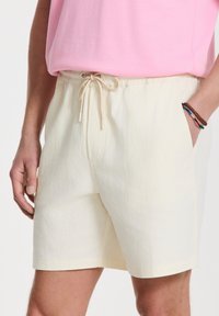Pantalones cortos de color beige claro con una textura suave, cinturilla elástica y cordón. Presenta bolsillos laterales y un diseño simple y minimalista.