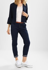 Marinblå peplumjacka över en vit ribbad topp, ihop med marinblå byxor och vita sneakers. Byxorna har blommigt detaljer i fållen. Brunt bälte ingår.