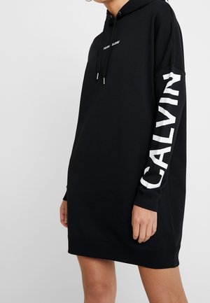Personne portant une robe à capuche noire avec "CALVIN" en grandes lettres blanches sur la manche droite et un petit texte blanc sur la poitrine.