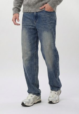 Osoba oblečená v sivom svetri, vybledlých modrých džínsoch a bielych teniskách New Balance, stojaca pred jednoduchým svetlým pozadím.