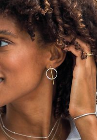 Orecchino lineare dorato con contorno circolare, caratterizzato da una barra verticale che si interseca. Indossato con collane delicate sovrapposte.