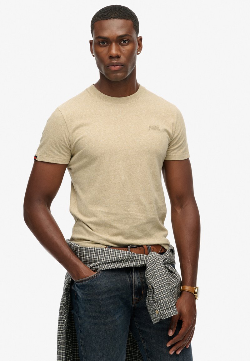 Uomo che indossa una t-shirt beige a maniche corte, jeans scuri, cintura marrone, orologio, orecchini, con una camicia a quadri legata in vita, in piedi davanti a uno sfondo bianco.