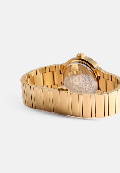 Reloj de metal dorado con un bisel decorado y una correa entrelazada. La pantalla cuenta con un fondo de cristal que muestra diseños intrincados.