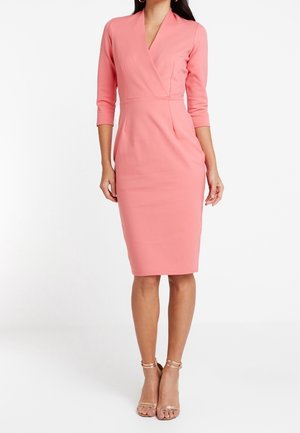 Kobieta w dopasowanej, różowej sukience do kolan z rękawami do połowy ramienia i dekoltem w serek, w połączeniu ze złotymi szpilkami na kostkę.