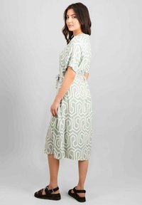 Abito verde con un motivo geometrico bianco, maniche corte, cintura e un design di lunghezza media. Indossato con sandali neri.
