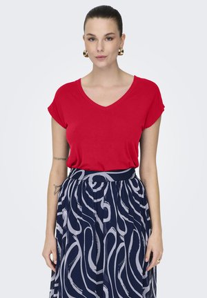 Mujer con blusa roja de manga corta y cuello en V, y falda azul marino de cintura alta con remolinos abstractos en gris claro, posando frente a un fondo liso.