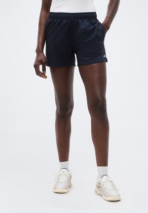 Osoba v čiernych športových šortkách, bielych ponožkách a bielych teniskách, stojaca pred jednoduchým svetlým pozadím.