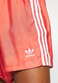 Korallinväriset juoksushortsit, joissa on valkoinen kolmiraitakuosi, sileä pinta ja valkoinen adidas-logo painettuna helman lähelle.