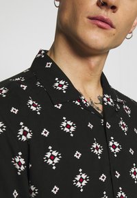 Zwart shirt met korte mouwen, voorzien van een herhalend geometrisch patroon in wit en rood, met een klassiek kraag en een gladde stofstructuur.