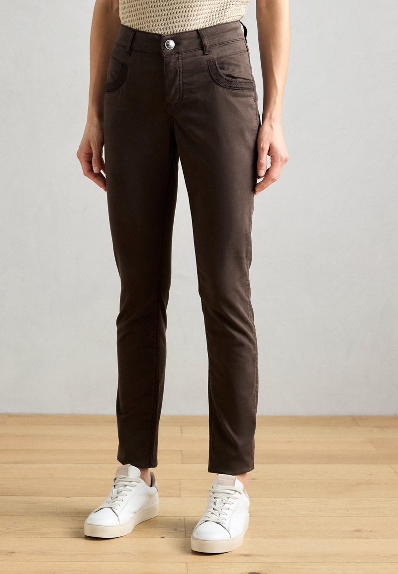Personne portant un pantalon slim marron foncé et des baskets blanches, debout sur un sol en bois contre un mur clair uni.