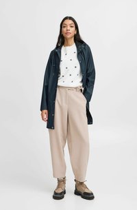Jachetă impermeabilă bleumarin, pulover alb cu model floral negru, pantaloni bej cu talie înaltă, și cizme împletite maronii cu tălpi negre.