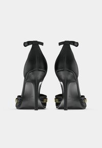 Crne kožne cipele na visoku petu s tankom petom i remenom oko gležnja. Imaju zlatne metalne detalje i glatku teksturu.