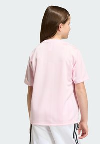 Jong persoon met lang bruin haar, gekleed in een lichtroze shirt met korte mouwen en witte sportshorts met zwarte strepen, van achteren gezien.