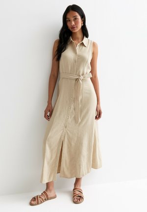 Abito maxi in lino beige con apertura a bottoni sul davanti, colletto e cintura in tessuto. Presenta uno spacco frontale ed è abbinato a sandali marroni con cinturini.