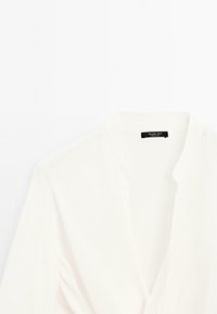 Blusa bianca con scollo a V e dettagli arricciati sotto il busto, caratterizzata da un colletto semplice e maniche lunghe.