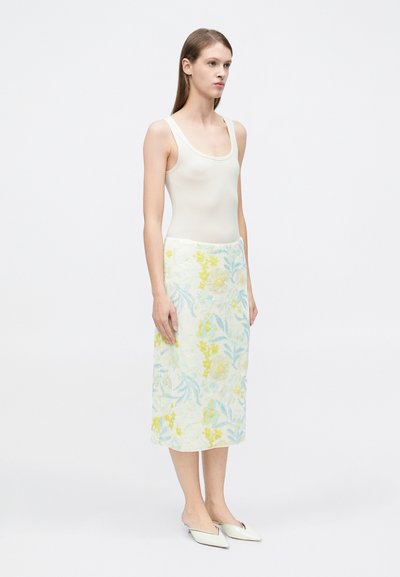 Jupe midi fleurie avec des motifs brodés jaunes et bleus, associée à un débardeur crème ajusté. Des chaussures plates blanches complètent le look.