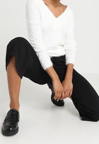 Pull blanc duveteux à encolure en V avec manches longues, associé à un pantalon court noir ample et des mocassins noirs texturés.