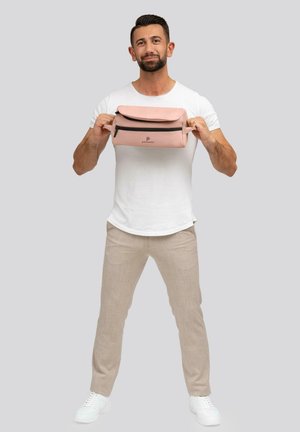 Sac en tissu rose doux avec un rabat pliable, fermeture éclair noire et logo. Le modèle porte une chemise blanche et un pantalon beige, tenant le sac au niveau de la poitrine.