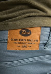 Vaqueros de denim azul claro con un parche de cuero marrón texturizado que presenta el logotipo de 'Petrol' y texto sobre la calidad de la artesanía.