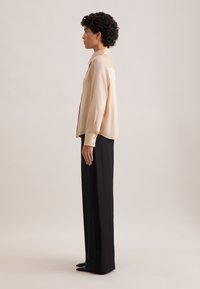 Blusa beige a maniche lunghe con colletto, caratterizzata da una texture setosa e dettagli con bottoni, abbinata a pantaloni neri a vita alta e scarpe a punta.