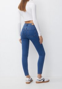 Mujer con jeans ajustados de cintura alta de color azul, top corto de manga larga blanca y zapatillas blancas con suelas de goma, de pie contra una pared blanca.